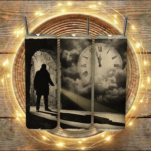 May include: Three stainless steel tumblers with black straws, each with a sepia-toned design. One shows a person in a doorway, another a clock, and the third a cloudy sky. The tumblers are arranged on a wooden surface.