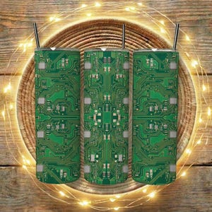 May include: Three green and white insulated tumblers with a circuit board design, each with a silver straw. The tumblers are arranged on a woven basket, with string lights and a wooden background.