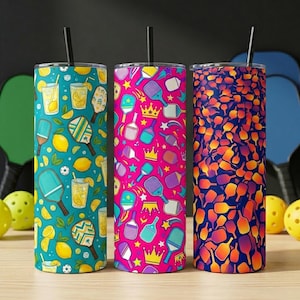 May include: Three cylindrical tumblers with straws, each featuring a distinct pattern. The left tumbler is teal with pickleball paddles, lemons, and drinks. The middle is pink with paddles and crowns. The right is a gradient of orange to blue with paddles.