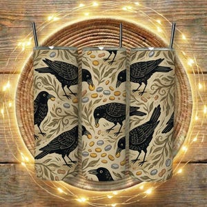 May include: Three stainless steel tumblers featuring black crow designs on a beige backdrop. The pattern includes peanuts and bottle caps, with a leafy motif. Each tumbler has a silver straw. The tumblers are displayed on a woven basket.