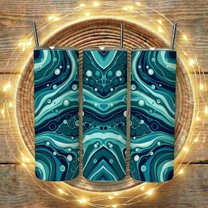 Op de afbeelding: Drie geïsoleerde bekers in teal en blauw met een wervelend abstract ontwerp, elk met een zilveren rietje. De bekers zijn gerangschikt op een geweven mand, met lichtslingers en een houten achtergrond.