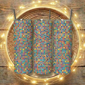 Puede incluir: Tres vasos de acero inoxidable con pajitas, cada uno cubierto con un patrón de mosaico de cuadrados coloridos. Un birrete de graduación y el año "2023" aparecen en el vaso central. Los vasos están colocados sobre un posavasos tejido, con luces de cadena.