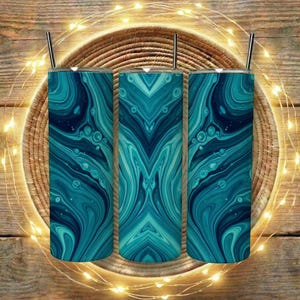 Op de afbeelding: Drie roestvrijstalen bekers met een abstract wervelontwerp in blauwgroen en marineblauw. Elke beker heeft een zilveren rietje en staat op een geweven mand, met lichtslingers rond de mand. De bekers staan op een houten oppervlak.