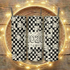 Peut inclure: Trois gobelets sur le thème de la remise des diplômes avec des motifs à carreaux noirs et blancs. Le design comprend des chapeaux de graduation, des diplômes et l'année "2026". Chaque gobelet a une paille argentée. Les gobelets sont posés sur un tapis tissé avec des guirlandes lumineuses.