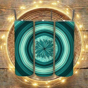 May include: Three teal-coloured tumblers with a radial design of concentric circles and a central geometric flower. Each tumbler has a black straw. The tumblers are on a woven tray, with string lights on a wooden surface.