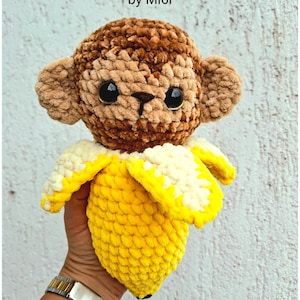 May include: A crochet toy depicting a monkey emerging from a banana. The monkey has brown fur, large black eyes, and tan ears. The banana is yellow with a white interior. The text "Chimpanzini Bananini" and "by Mi6i" are at the top of the image.