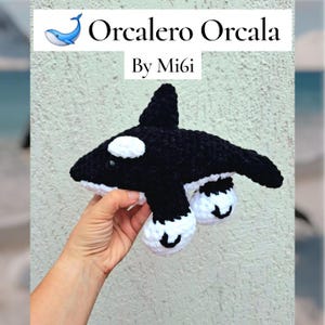 May include: A handmade, black and white crochet orca whale toy. The toy has white patches, a dorsal fin, and small, round eyes. The text "Orcalero Orcala By Mi6i" is displayed at the top of the image.