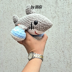May include: A crocheted shark toy in shades of gray, with black eyes, a red mouth, and blue and white sneakers. The text "Tralalelotralala the Shark with Sneakers" and "by Mi6i" are visible.