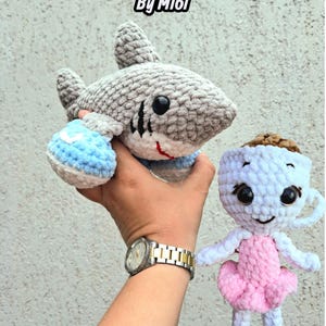May include: A bundle of two handmade crochet toys: a gray shark with black accents and a red mouth, and a doll in a pink tutu. The shark has blue and white fins. The text "BUNDLE By Mi6i" is at the top.