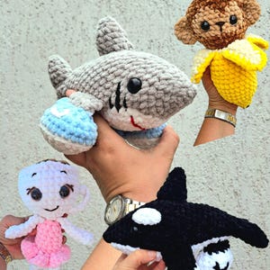 Peut inclure: Ensemble de quatre jouets amigurumi crochetés à la main : un requin, un singe dans une banane, une poupée tasse à thé et une orque. Les jouets sont de couleurs variées, dont le gris, le marron, le jaune, le rose, le blanc et le noir. Le texte "BRAINROT CROCHET PATTERN BUNDLE - 4 AMIGURUMI TOYS PATTERN" est en haut.