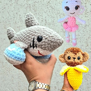 May include: Crocheted plush toys: a gray shark with blue accents, a ballerina doll in pink, and a monkey with a yellow banana. The shark has black stripes and a red mouth. The toys are soft and handmade.