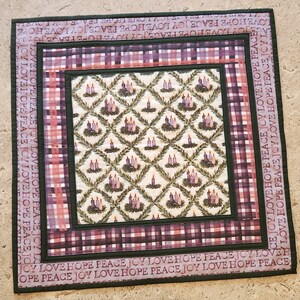 May include: A quilted table runner with a green, white, and purple plaid pattern. The center of the runner features a repeating pattern of candles and greenery. The border of the runner has the words "Joy Love Hope Peace" repeated in a dark green font.