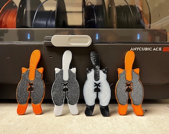 May include: Four cat butt-shaped 3D-printed objects in various colour combinations: orange and grey, white and grey, black and white, and orange and grey. The objects are displayed in front of a 3D printer.