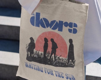 May include: A beige canvas tote bag with the band name "The Doors" in blue stencil font. Below is a silhouette of the band members in front of a red sun, with the text "Waiting for the Sun". The bag is held by a person.