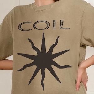 May include: Khaki-coloured t-shirt featuring the word "COIL" and a black sunburst design. The shirt has a relaxed fit and is made of a soft material. The t-shirt is a neutral colour.