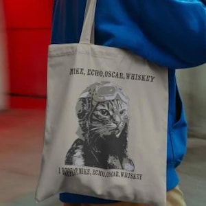 May include: Beige tote bag featuring a black and white illustration of a cat wearing a pilot's helmet. The text "MIKE, ECHO, OSCAR, WHISKEY" is printed above the image, and below it, the same text is repeated.