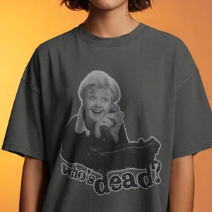 Puede incluir: Una camiseta de manga corta en color gris carbón con un estampado gráfico de una persona sosteniendo un auricular de teléfono. Debajo de la figura, el texto "who's dead?" está impreso en una fuente redondeada y estilizada. Cuello redondo y corte holgado.