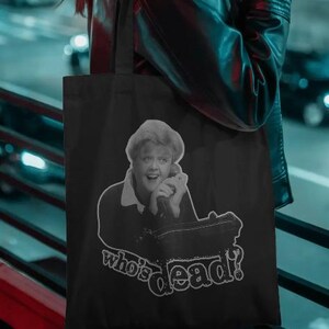 Puede incluir: Bolso tote negro con una imagen en blanco y negro de una mujer al teléfono y el texto "who's dead?" en una fuente estilizada. El bolso lo lleva una persona con una chaqueta de cuero negra, con un fondo de ciudad.