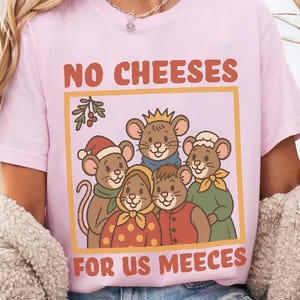 May include: Light pink t-shirt featuring a cartoon illustration of five mice wearing various outfits, with the text "NO CHEESES FOR US MEEECES" in brown letters. The design is framed in a yellow rectangle. The shirt is a casual wear item.