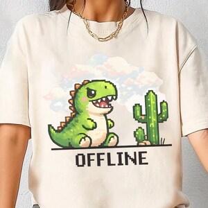 Pode incluir: Camiseta cor marfim com um gráfico pixelado de um dinossauro verde e um cacto, com a palavra "OFFLINE" em preto. O dinossauro tem uma expressão amigável. A camiseta tem um corte casual e oversized.