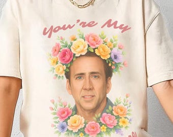 May include: Ivory-colored t-shirt featuring a portrait of Nicolas Cage adorned with a floral crown and the text "You're My National Treasure." The design includes pink, yellow, and purple flowers. The shirt has short sleeves.