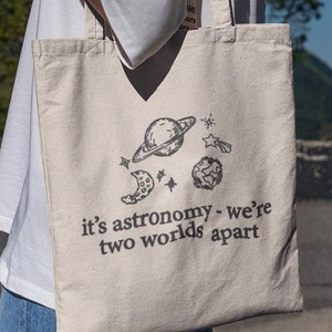 Könnte beinhalten: Eine beige Tragetasche mit einer schwarz-weißen Illustration von Planeten und Sternen. Die Tasche trägt den Text "it's astronomy - we're two worlds apart".