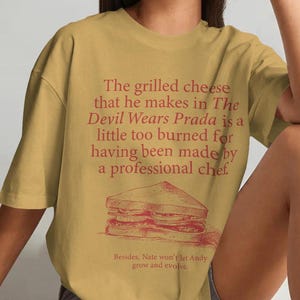 May include: A mustard yellow short-sleeve t-shirt featuring red text that reads, The grilled cheese that he makes in The Devil Wears Prada is a little too burned for having been made by a professional chef, with a graphic of a sandwich below.