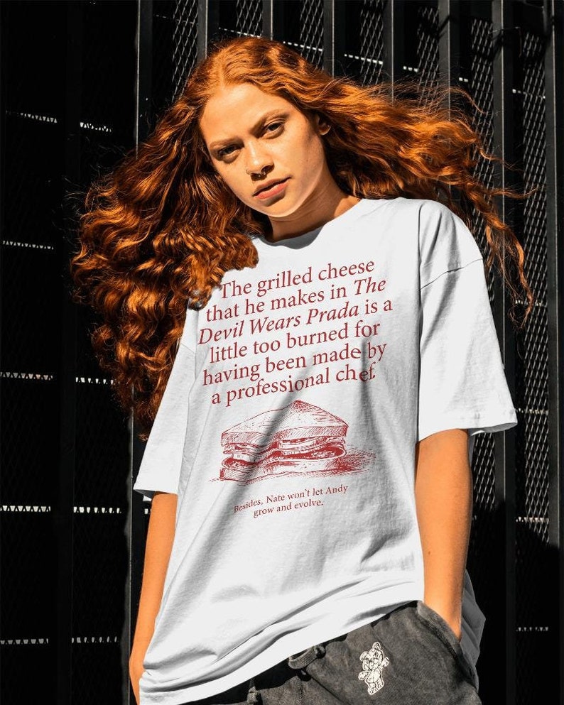 May include: White t-shirt with red text that reads "The grilled cheese that he makes in The Devil Wears Prada is a little too burned for having been made by a professional chef."  The text is accompanied by a black and white illustration of a grilled cheese sandwich.