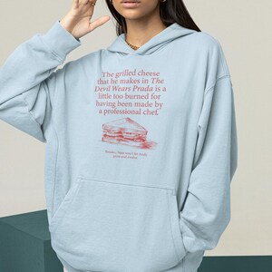 May include: Light blue hooded sweatshirt with a graphic print of a grilled cheese sandwich and the text "The grilled cheese that he makes in The Devil Wears Prada is a little too burned for having been made by a professional chef. Besides, Nate won't let Andy grow and evolve."