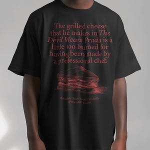 May include: Black t-shirt with a red graphic print of a grilled cheese sandwich and text that reads "The grilled cheese that he makes in The Devil Wears Prada is a little too burned for having been made by a professional chef. Besides, Nate won't let Andy grow and evolve."