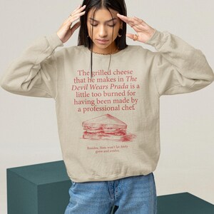 May include: A beige sweatshirt with a red graphic print of a grilled cheese sandwich and the text "The grilled cheese that he makes in The Devil Wears Prada is a little too burned for having been made by a professional chef' Besides. Nate won't let Andy grow and evolve."