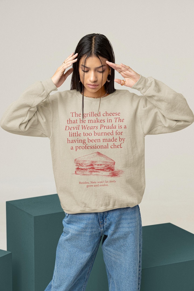 May include: A beige sweatshirt with a red graphic print of a grilled cheese sandwich and the text "The grilled cheese that he makes in The Devil Wears Prada is a little too burned for having been made by a professional chef' Besides. Nate won't let Andy grow and evolve."