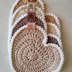 May include: A stack of handmade crochet coasters in shades of beige, brown, and white. Each coaster has a scalloped edge and a small leather tag that says "handmade". The coasters are arranged in a fan shape.