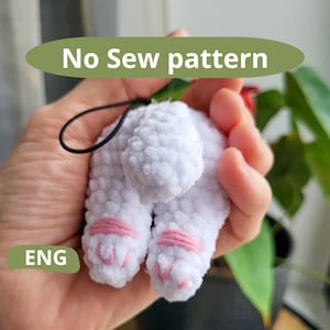 Op de afbeelding: Wit gebreid pluche speelgoed met roze accenten, gevormd als een klein dier met een staart. Het speelgoed heeft een zwarte lus om op te hangen. De tekst "No Sew pattern" is zichtbaar in de afbeelding.