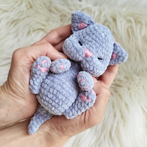Può includere: Un piccolo gatto di peluche lavorato all'uncinetto a mano con un morbido filato grigio chiaro. Il gatto presenta dettagli ricamati in rosa sulle orecchie, sul naso e sui cuscinetti delle zampe. Ha una testa arrotondata e un corpo paffuto.