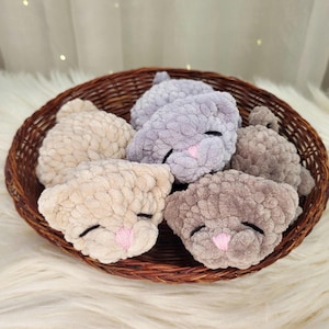 May include: A woven basket holds several plush, crocheted cat faces in various neutral colors. Each cat face has closed black eyes and a pink nose. The basket is brown and sits on a white, fluffy surface.