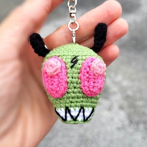 May include: Crocheted keychain featuring a green alien head with pink eyes, black antennae, and a white-toothed grin. The handmade keychain has a silver metal ring for attaching to keys or bags. The alien has a lightning bolt on its forehead.