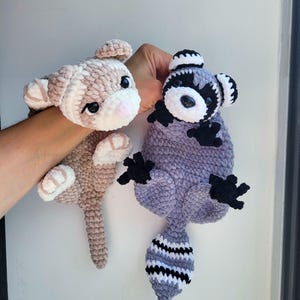 Op de afbeelding: Twee handgemaakte gehaakte knuffels: een lichtbruin en wit konijn en een grijze, zwarte en witte wasbeer. Het konijn heeft een roze neus en de wasbeer heeft een gestreepte staart. Beide zijn zacht en pluche.