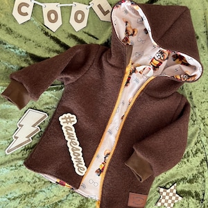 Könnte beinhalten: Braune Kapuzenjacke mit gelbem Reißverschluss und einem Innenfutter mit Cartoon-Motiven. Die Jacke hat braune Bündchen und einen kleinen Aufnäher. Holzausschnitte buchstabieren "COOL" und ein Blitz und der Hashtag "#awesome" sind ebenfalls sichtbar.