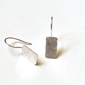 May include: A pair of silver-toned earrings featuring rectangular pendants with a hammered, textured surface. Each pendant is attached to a thin, curved wire hook designed for insertion into the earlobe, creating a simple drop style.