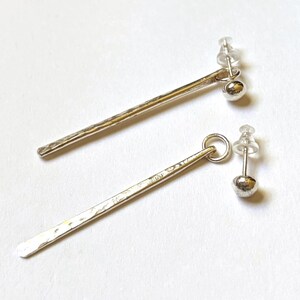 May include: A pair of silver-toned earrings featuring a long, thin, hammered metal bar suspended from a small circular ring attached to a spherical stud post with a clear plastic backing. The metal surface has a textured, uneven finish.