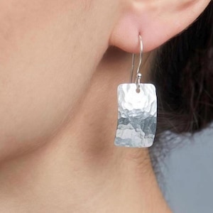 May include: A silver drop earring featuring a rectangular pendant with a textured, hammered finish. The pendant hangs from a thin, curved silver wire hook that passes through the earlobe, creating a simple and reflective metallic accessory.