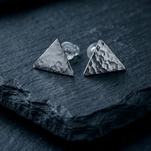 May include: A pair of silver-toned stud earrings shaped as triangles with a hammered, textured surface. Each earring features a pointed triangular face and includes a clear, translucent plastic backing for secure attachment to the earlobe.