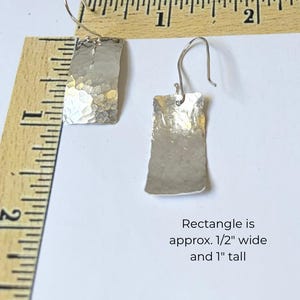 May include: Two silver-toned earrings with hammered rectangular drops and curved ear wires. A wooden ruler shows the earrings are approximately 0.5 inches wide and 1 inch tall, as noted by the text: Rectangle is approx. 1/2" wide and 1" tall.