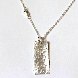 May include: A silver-colored necklace featuring a rectangular pendant with a textured, hammered finish. The pendant hangs from a delicate, thin silver-colored chain that includes a small circular jump ring and a lobster claw clasp closure.