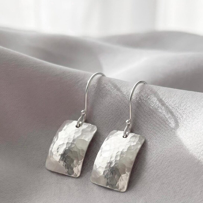 May include: A pair of silver drop earrings featuring rectangular, hammered metal pendants with curved edges and hook-style findings. The text "Handcrafted to last a lifetime." is displayed above the earrings in a black serif font.