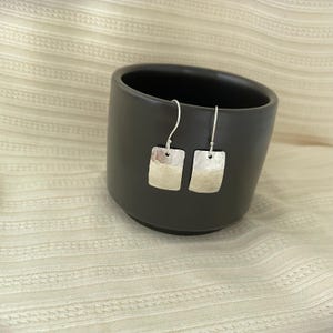 May include: A pair of silver drop earrings featuring rectangular pendants with a hammered texture. The earrings are suspended from thin silver ear wires and have a polished finish that reflects light across the metal surface.