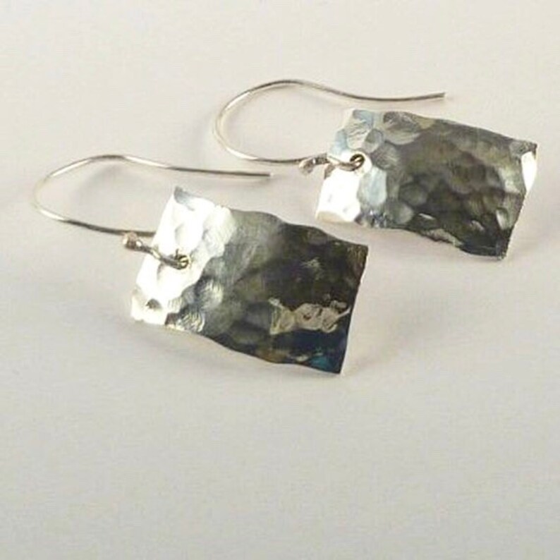 May include: A pair of silver-toned drop earrings featuring rectangular metal pendants with a hammered, textured surface. Each pendant is suspended from a curved silver-toned ear wire, creating a simple and reflective metallic design.