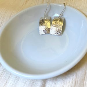 May include: A pair of silver-colored drop earrings featuring rectangular, slightly curved metal plates with a hammered, textured finish. The earrings are attached to simple silver-colored ear wires that hook through the top of each metal piece.
