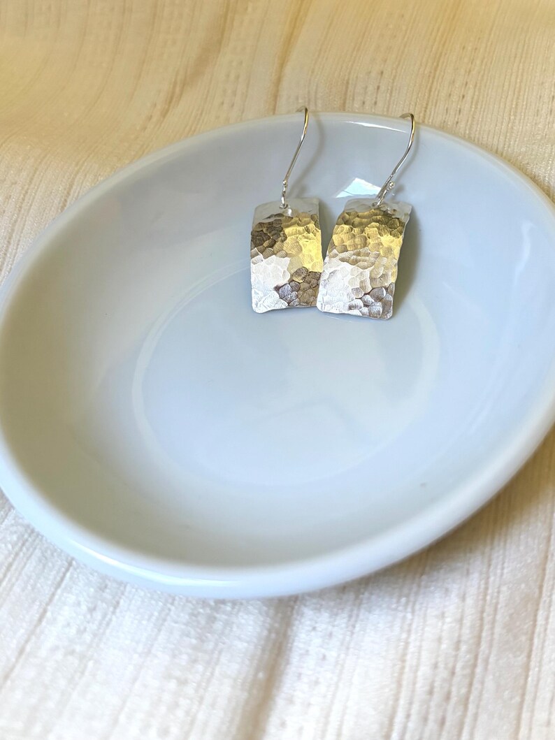 May include: A pair of silver-colored drop earrings featuring rectangular, slightly curved metal plates with a hammered, textured finish. The earrings are attached to simple silver-colored ear wires that hook through the top of each metal piece.